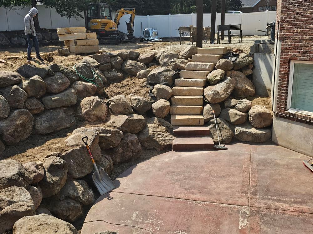 Landscape construction site featuring stone steps, rocks, and garden tools in a residential yard.