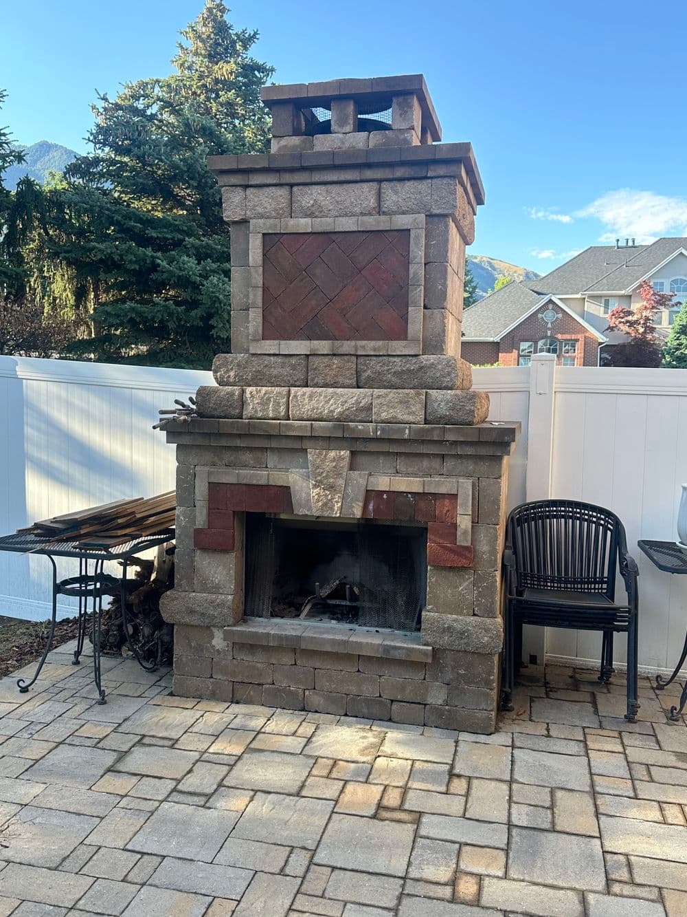 Outdoor stone chimney fireplace with patio, surrounded by trees and a white fence.