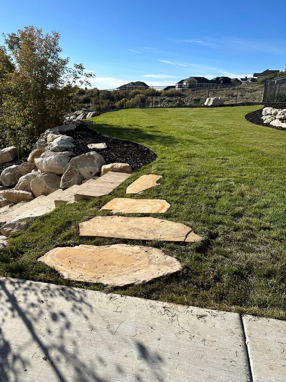 Stone pathway through landscaped yard with grass and rock features under a clear blue sky.